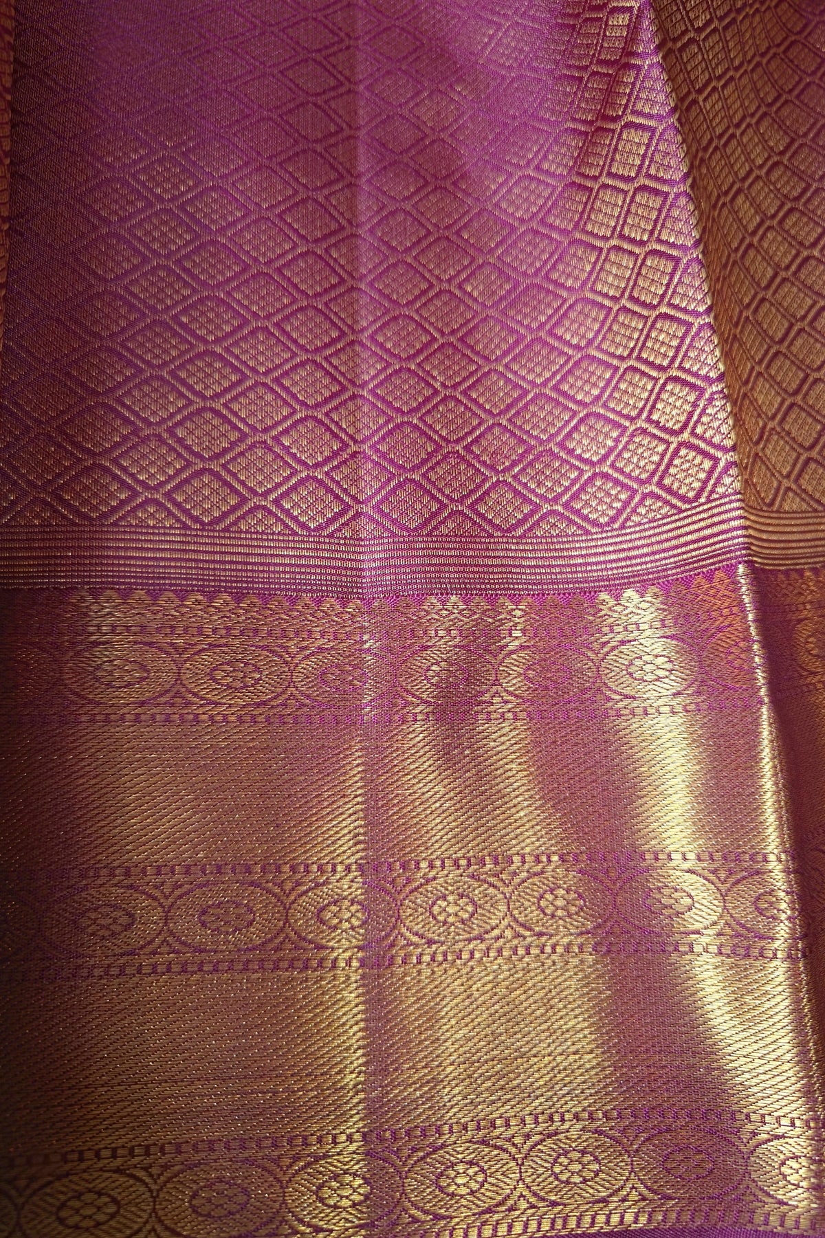 A close-up of a purple and gold woven fabric, likely a silk saree or textile, featuring intricate metallic zari patterns and a rich brocade border.