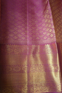 A close-up of a purple and gold woven fabric, likely a silk saree or textile, featuring intricate metallic zari patterns and a rich brocade border.