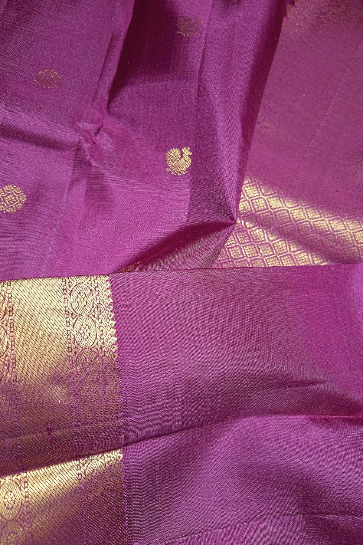 A close-up of a magenta purple silk saree with a wide gold zari border and small gold motifs woven into the fabric.