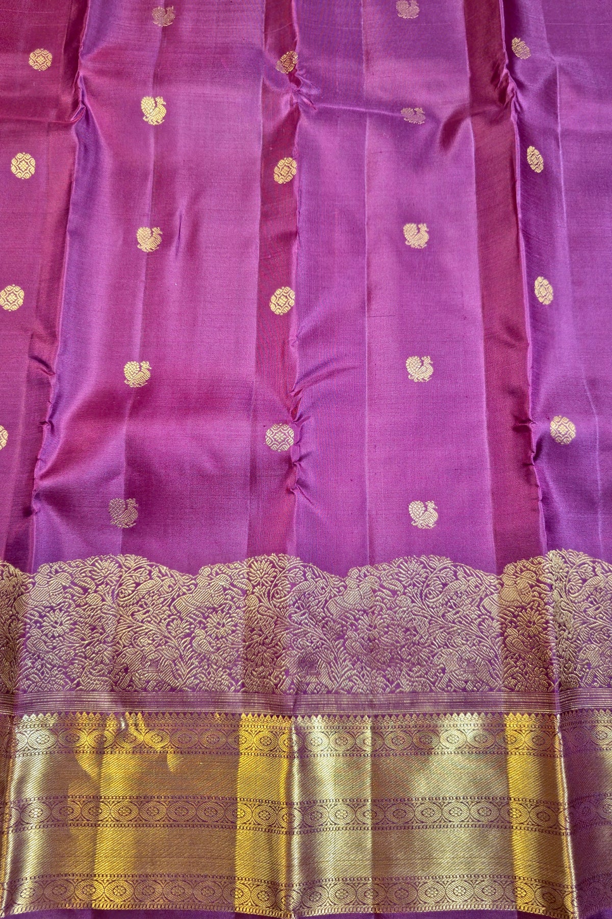 A close-up of a purple silk fabric, likely a Banarasi saree, featuring small gold zari floral motifs and a wide ornate gold zari border with a scalloped edge.