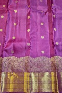 A close-up of a purple silk fabric, likely a Banarasi saree, featuring small gold zari floral motifs and a wide ornate gold zari border with a scalloped edge.