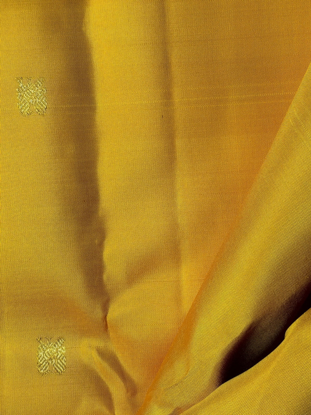 A close-up of a yellow fabric with a subtle sheen, showing soft folds and two small metallic embroidered motifs near the left side.