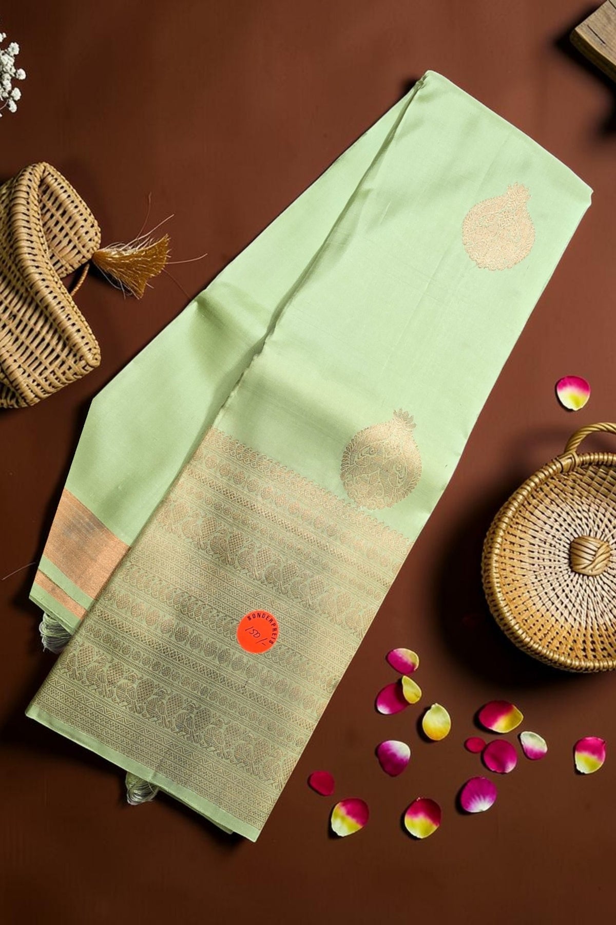 Light green saree with gold patterns on a brown surface with wicker basket and rose petals.