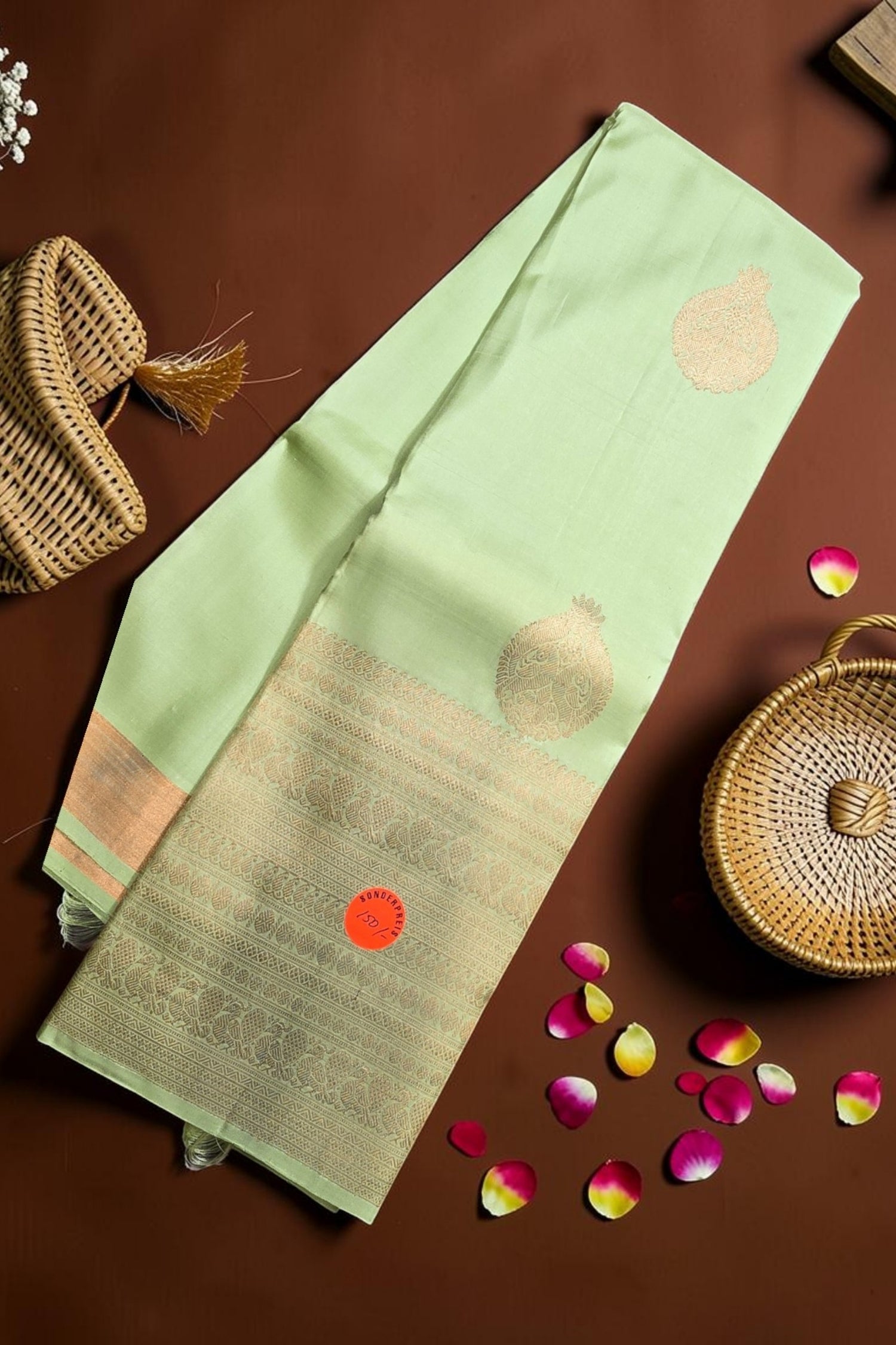 Light green saree with gold patterns on a brown surface with wicker basket and rose petals.