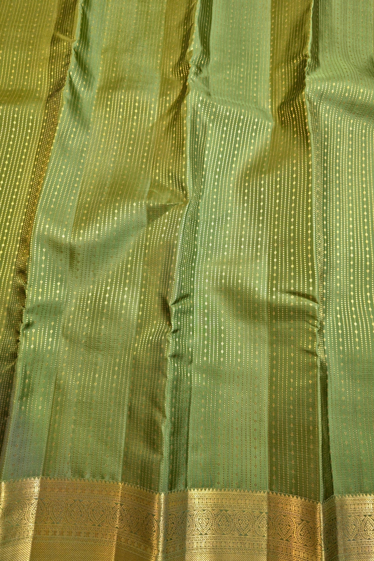 A close-up of a green silk fabric, likely a sari, featuring vertical stripes of small gold dots and a wide gold zari border at the bottom.