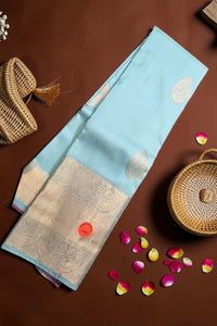 Light blue saree with gold patterns on a brown surface with a woven basket and rose petals.