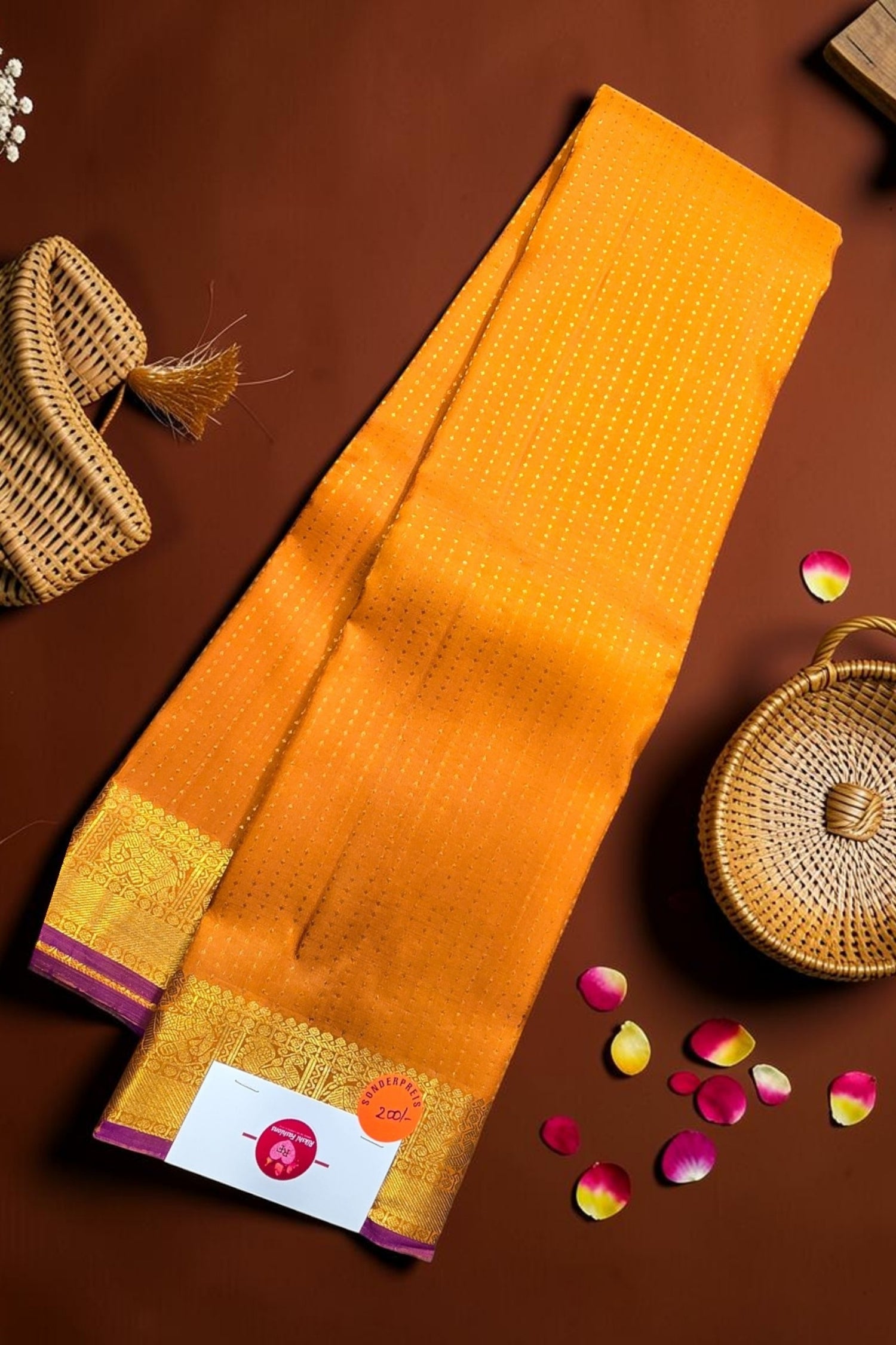 Yellow saree with a brand label on a brown background with baskets and petals.