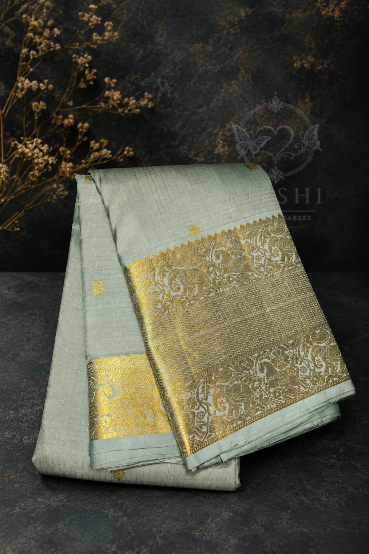 A folded light sage green silk saree with a wide, ornate gold zari border, displayed on a dark textured surface with dried floral props.
