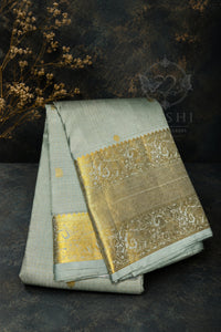 A folded light sage green silk saree with a wide, ornate gold zari border, displayed on a dark textured surface with dried floral props.