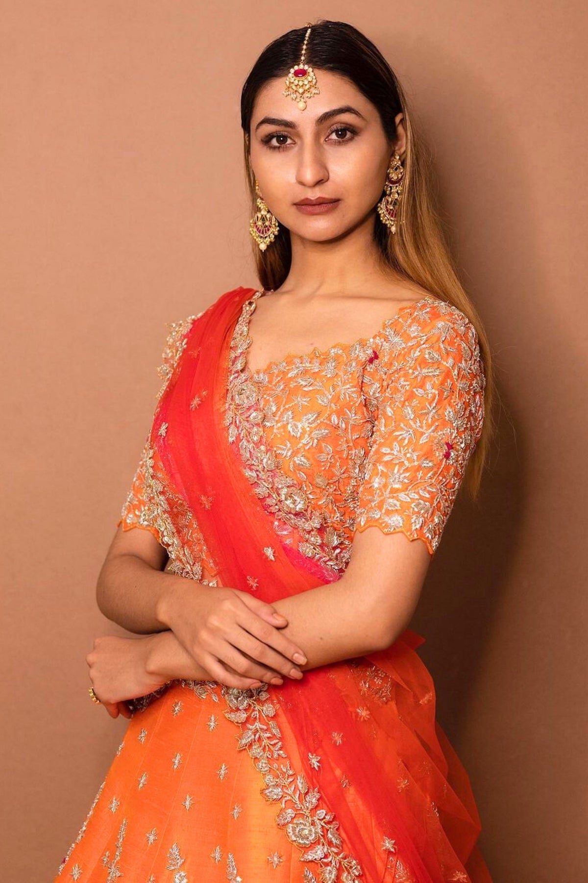 Woman wearing an orange and red traditional outfit with intricate designs against a brown background