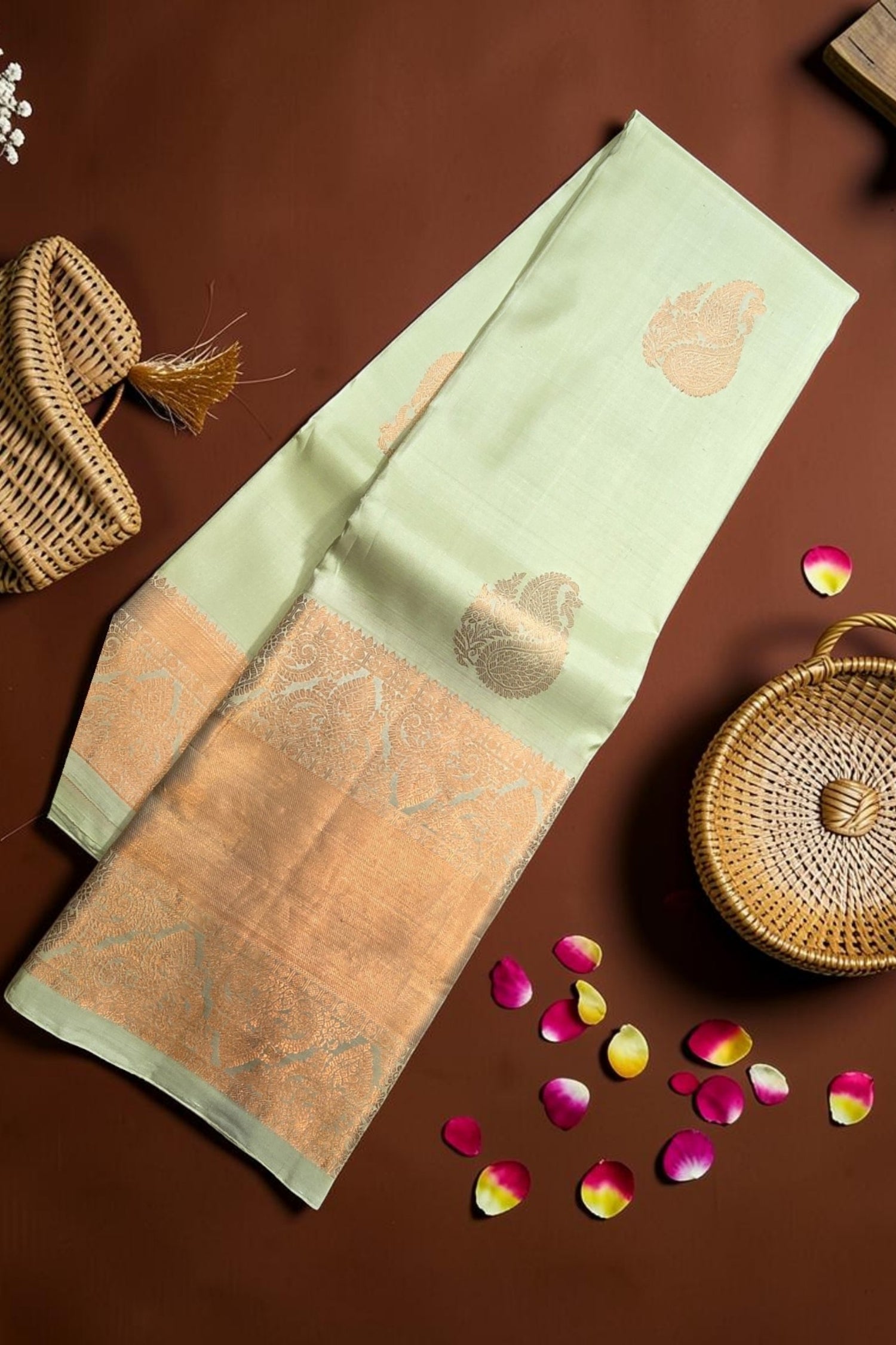 Light green fabric with gold patterns on a brown surface with wicker baskets and rose petals.