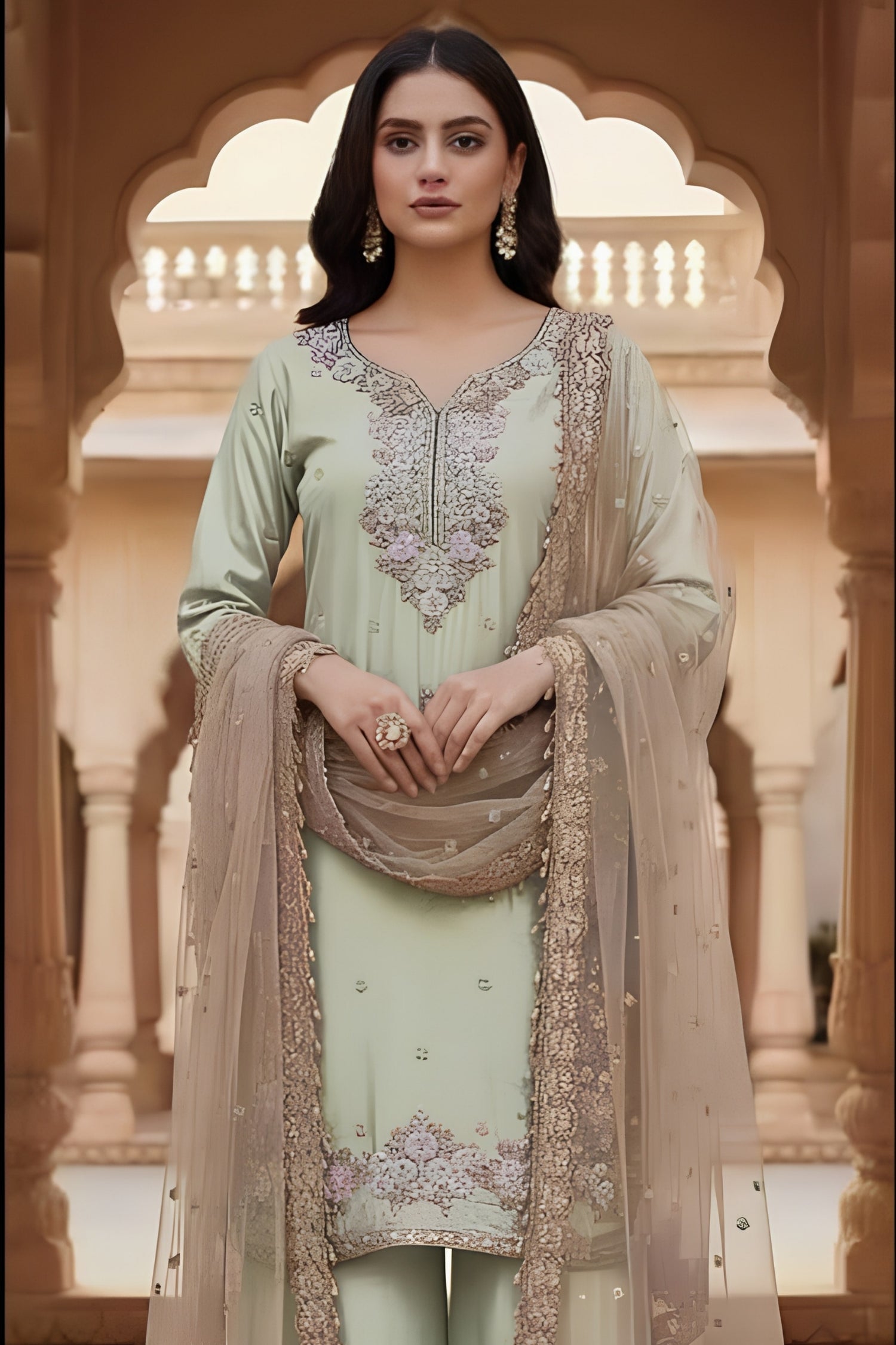 Woman in a light green traditional outfit with intricate embroidery, standing in front of an architectural background.
