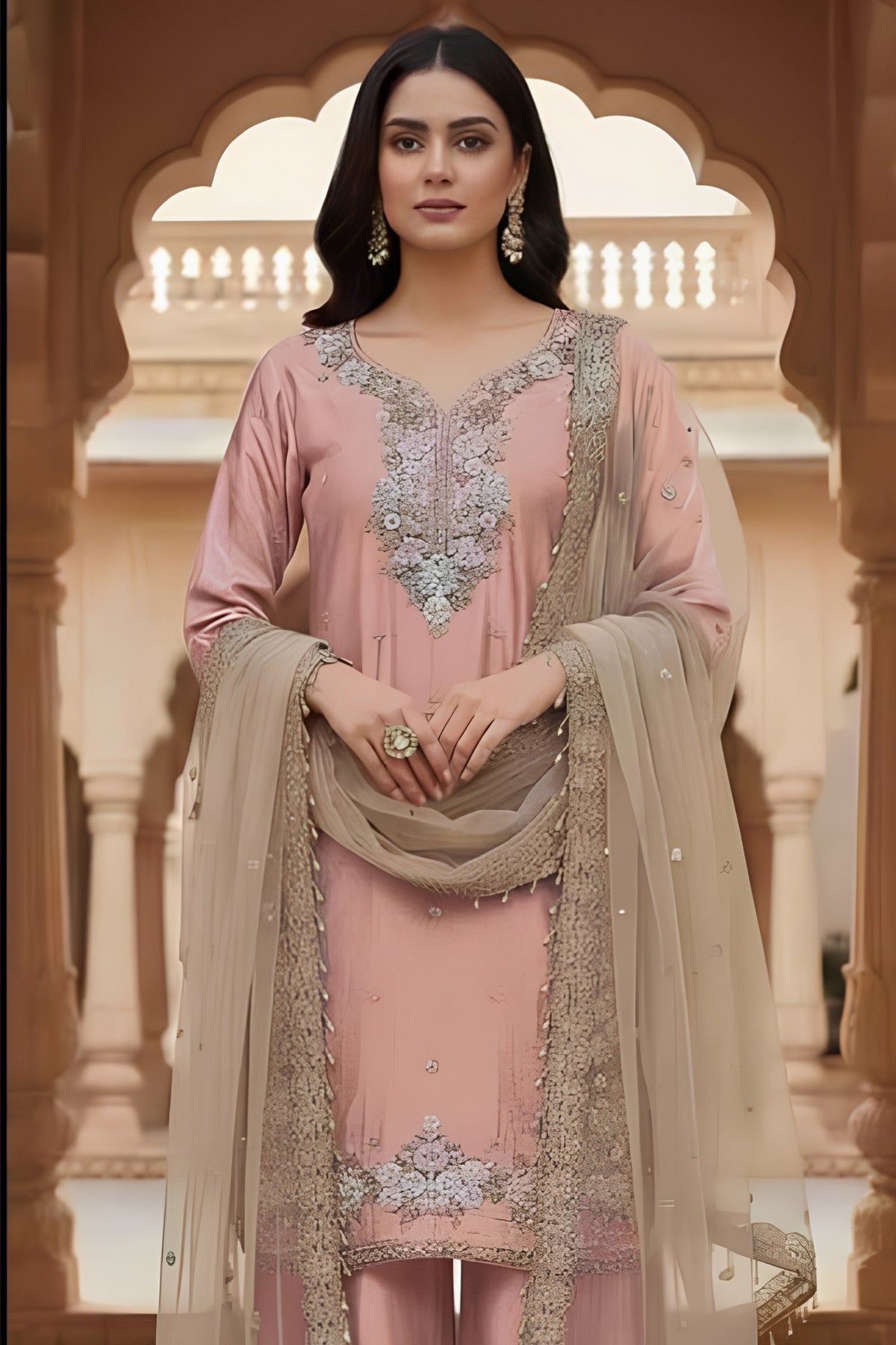 Woman in a traditional pink and beige outfit with intricate designs, standing in an ornate architectural setting.