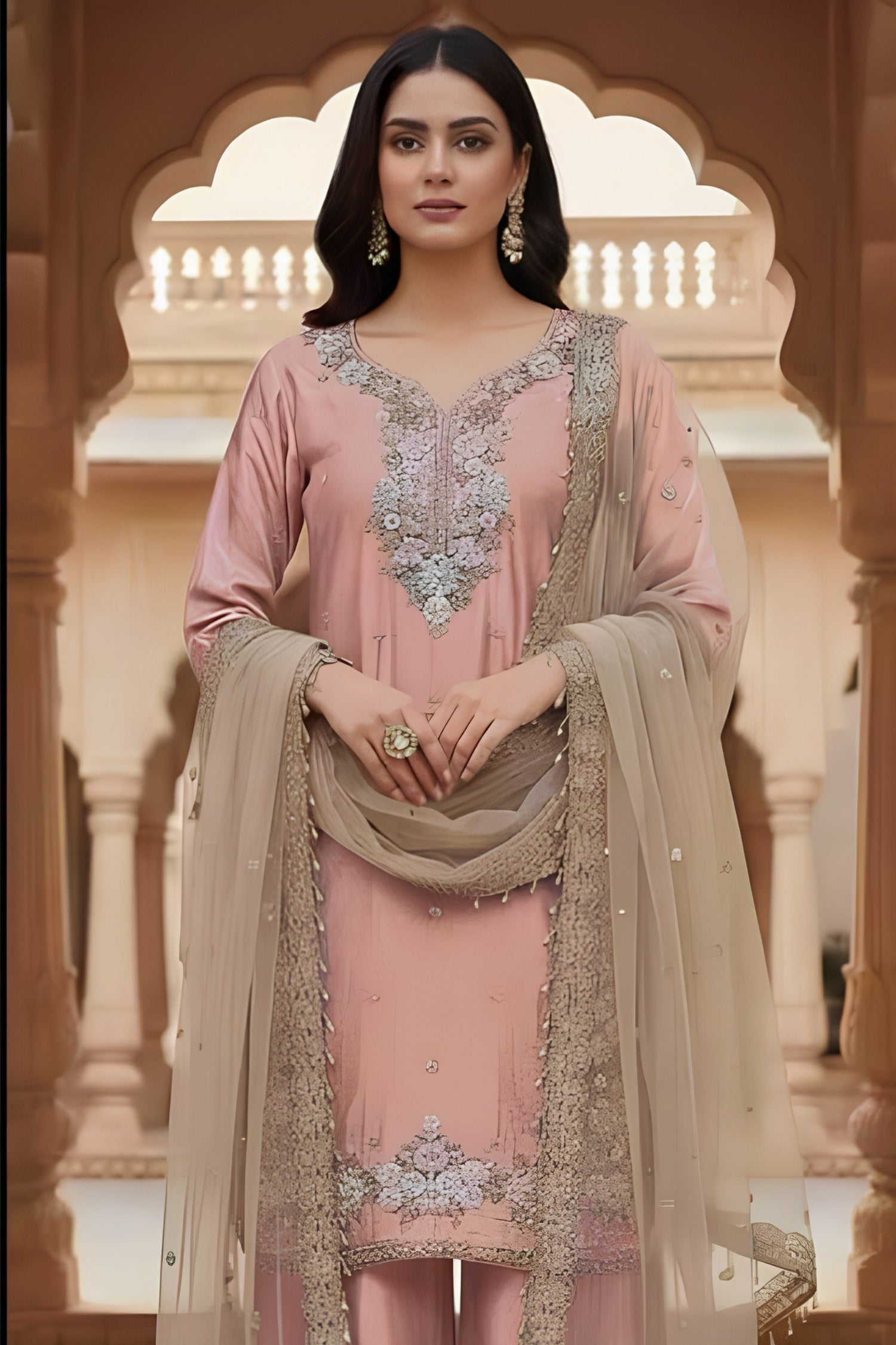 Woman in a traditional pink and beige outfit with intricate designs, standing in an ornate architectural setting.