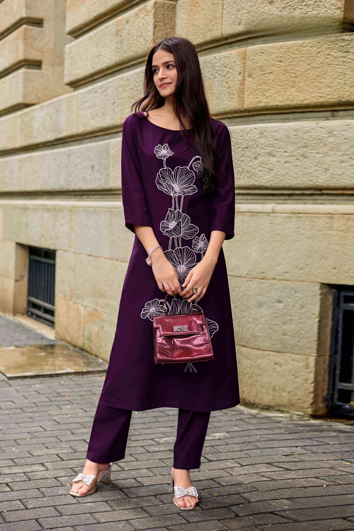 Woman in a purple outfit holding a matching handbag against a stone building background