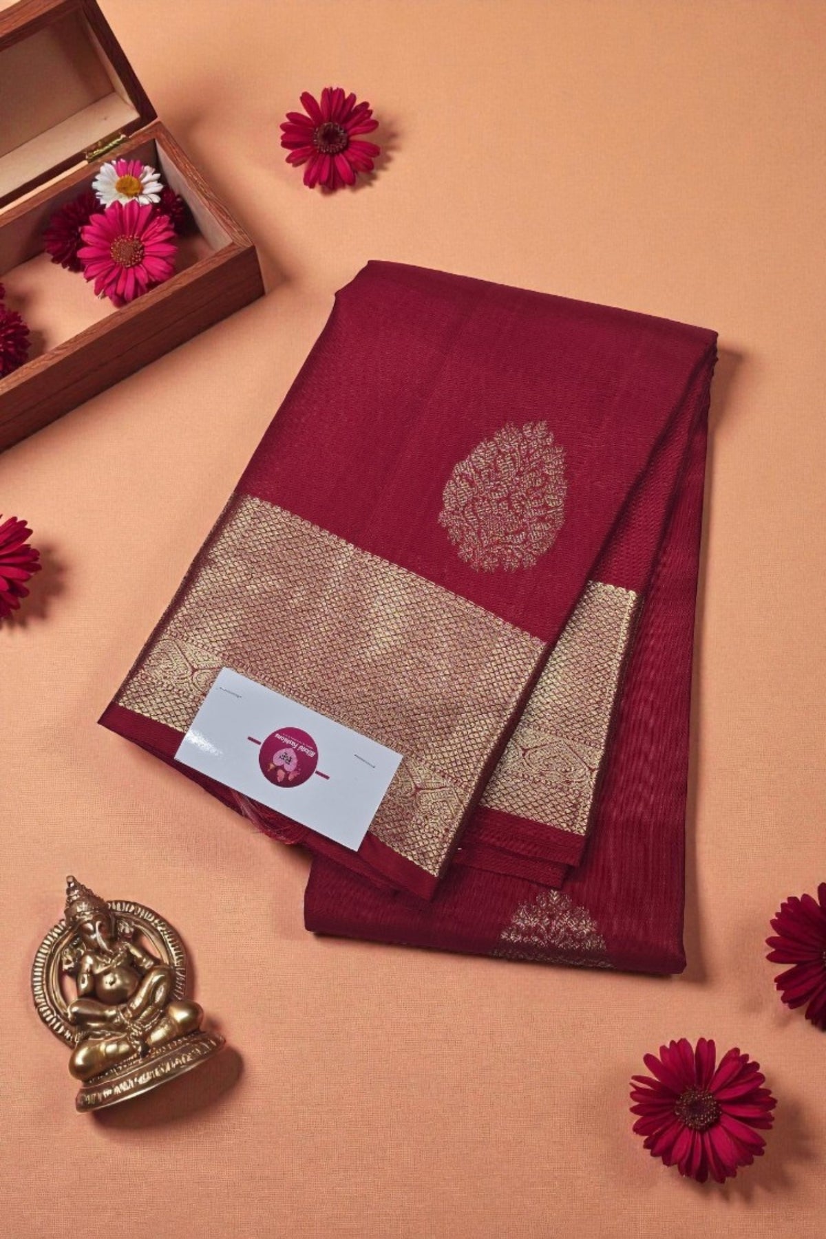 Maroon saree with gold border on a peach background with flowers and a decorative item.