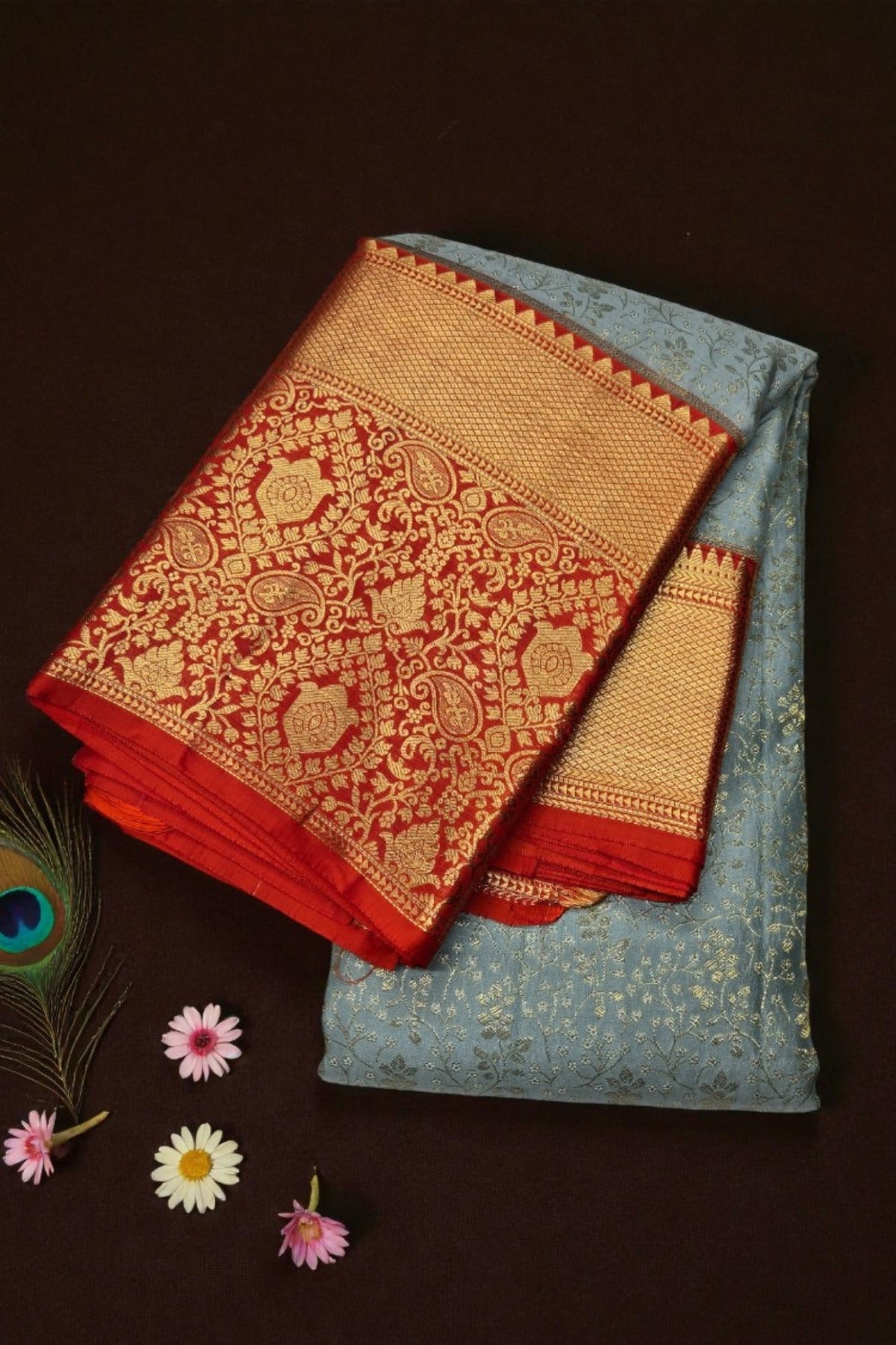 Two folded sarees, one red with gold patterns and the other blue with gold patterns, on a dark background.