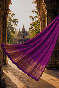 A flowing purple fabric with gold woven borders and small gold motifs, draped in the foreground between stone arches, with a temple and trees visible in the background.