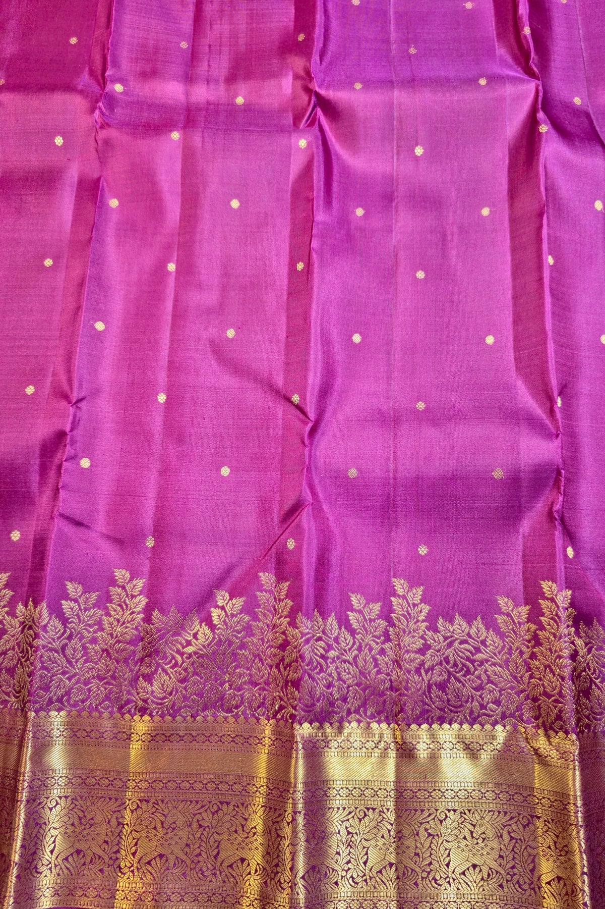 A close-up of a purple silk fabric, likely a sari, featuring small gold dot motifs and a wide ornate gold zari border at the bottom.