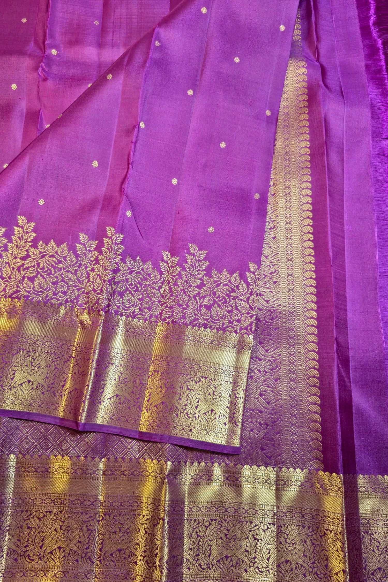 A close-up of a purple silk saree with gold zari embroidery, featuring a wide decorative border and small gold dots on the fabric.