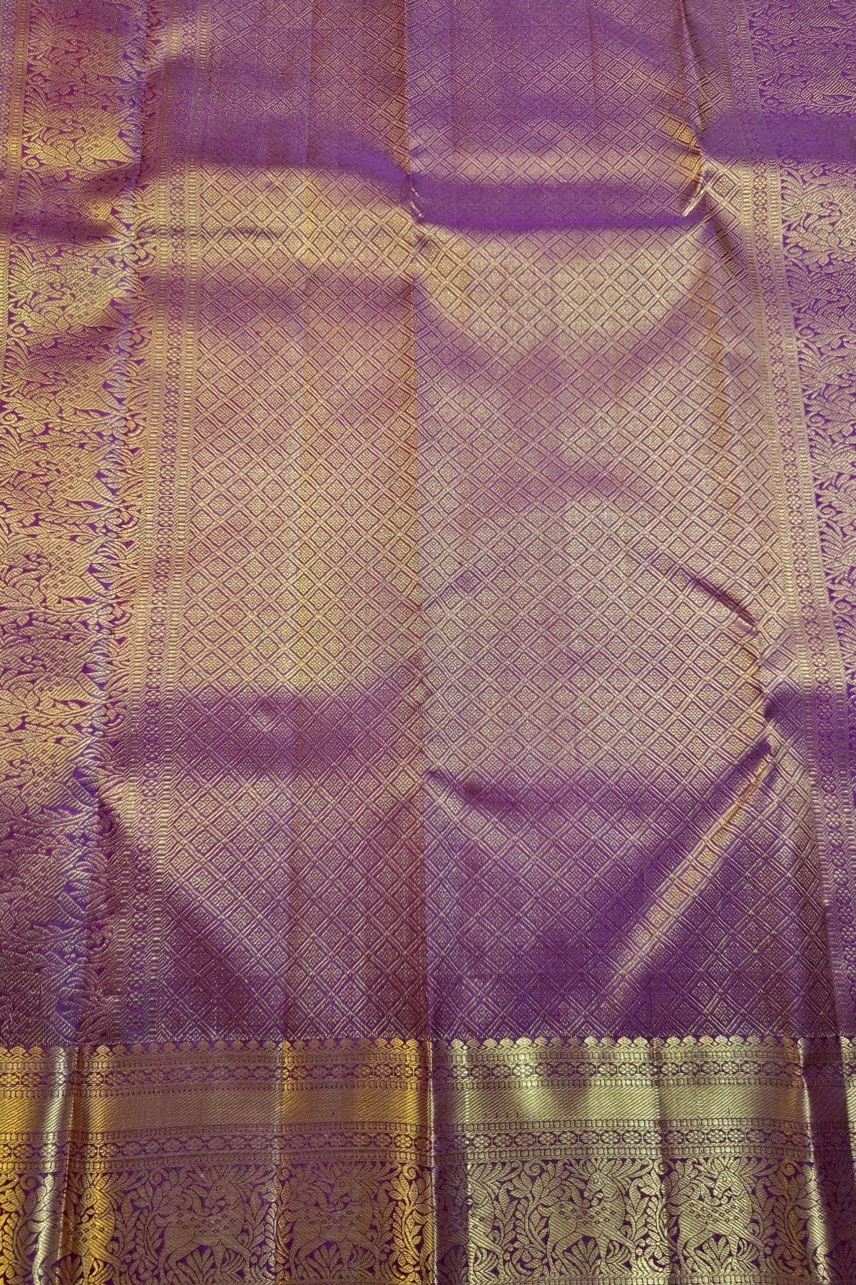 A close-up of a purple silk fabric with intricate gold zari woven patterns and a decorative border.