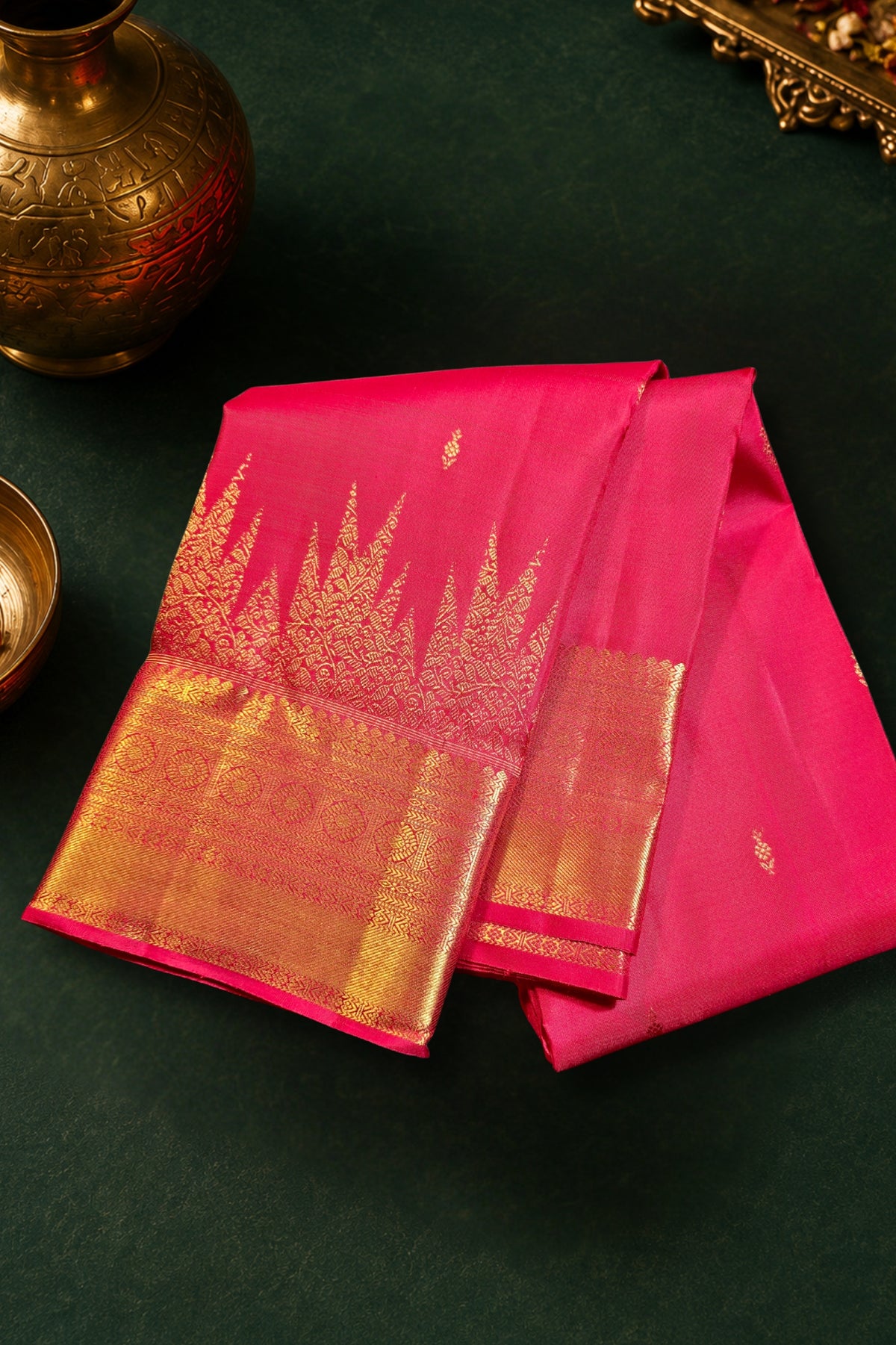 A folded pink silk saree with a wide gold zari border and floral motifs, displayed on a dark green textured surface with traditional brass decorative props.
