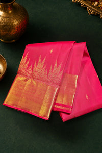 A folded pink silk saree with a wide gold zari border and floral motifs, displayed on a dark green textured surface with traditional brass decorative props.