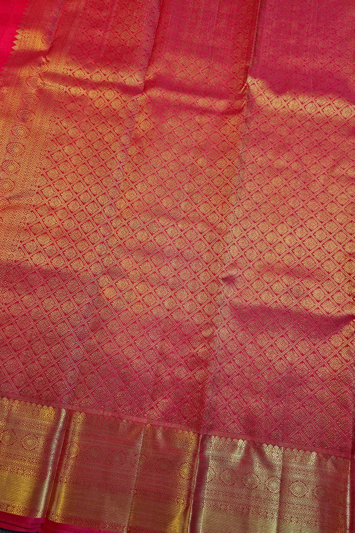 A close-up of a pink and gold woven fabric, likely a sari, featuring a repeating geometric pattern and a wide metallic gold border.