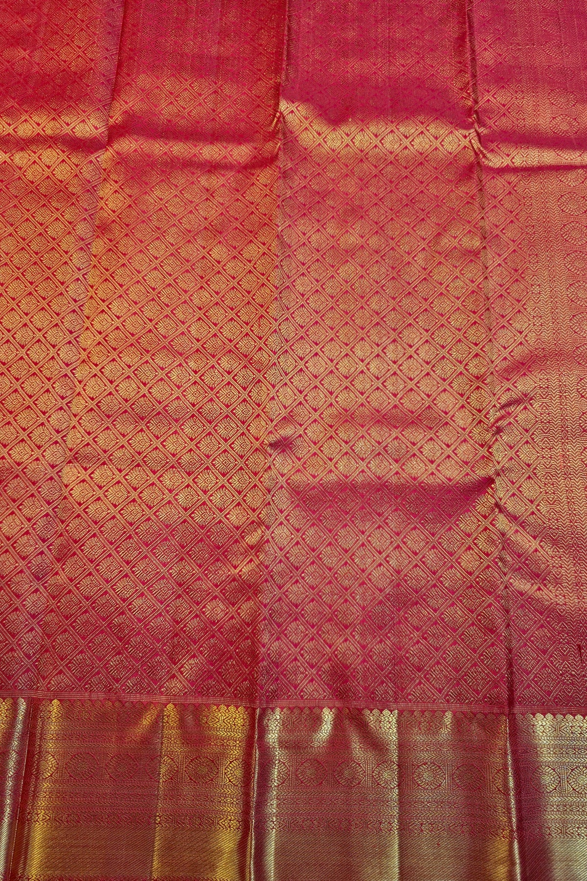 A close-up of a red and gold woven fabric, likely a silk saree or textile, featuring a detailed gold zari brocade pattern and a decorative gold border at the bottom.