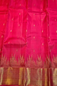 A close-up of a bright pink silk saree or fabric with small gold motifs and a wide gold zari border along the bottom edge.