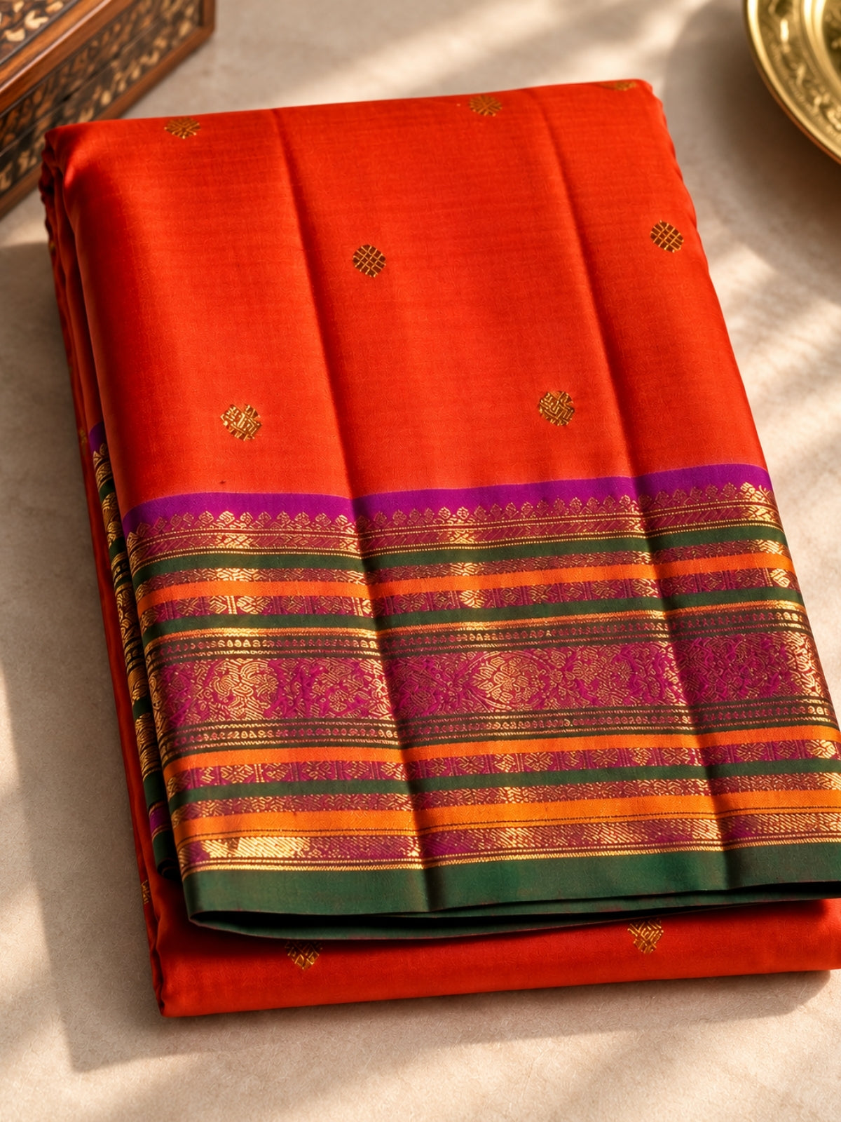 A folded orange-red silk saree with a wide multicolored zari border and small gold motifs, displayed on a light fabric surface.