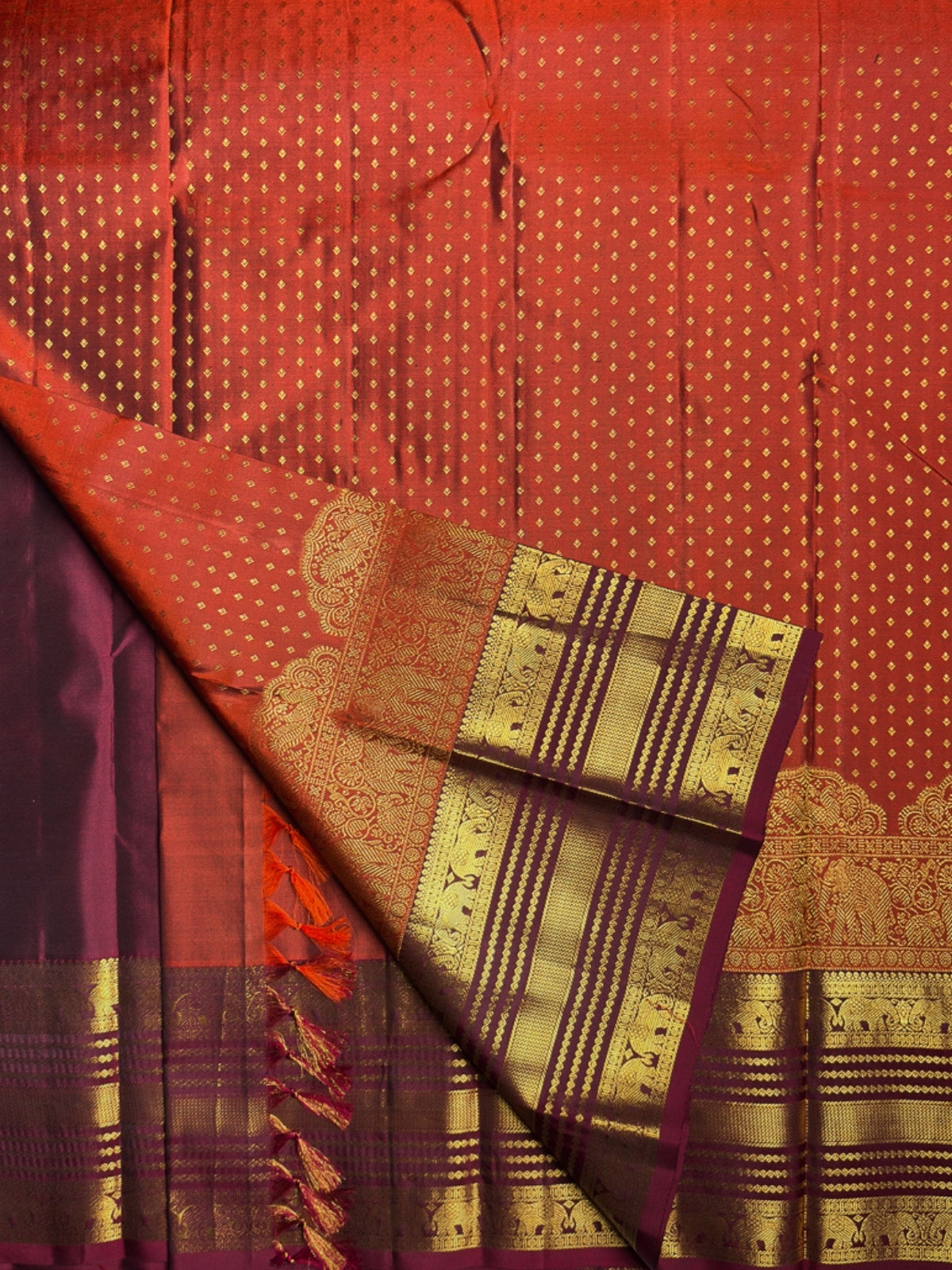 A close-up of a red and deep purple silk saree with intricate gold zari woven patterns, borders, and tassels.