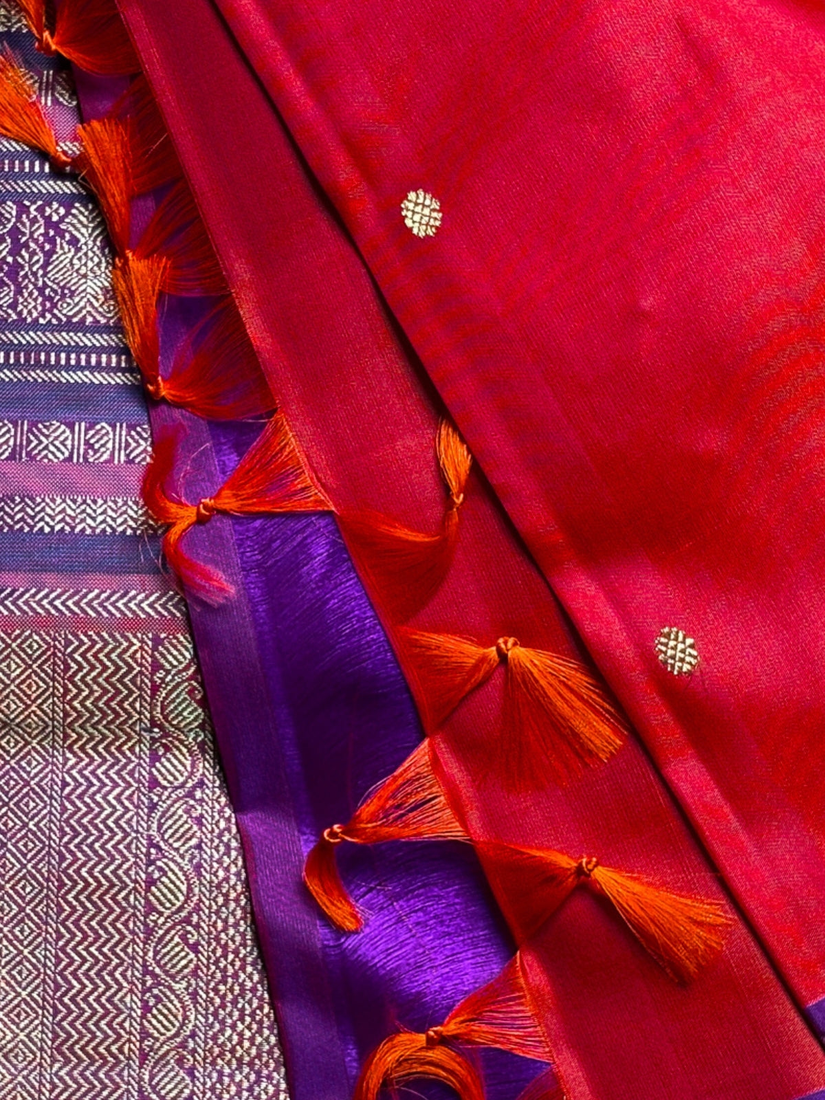 A close-up of a richly colored fabric, likely a sari, featuring a dominant bright red body with orange tassels and small gold motifs, layered over a purple and gold patterned border.