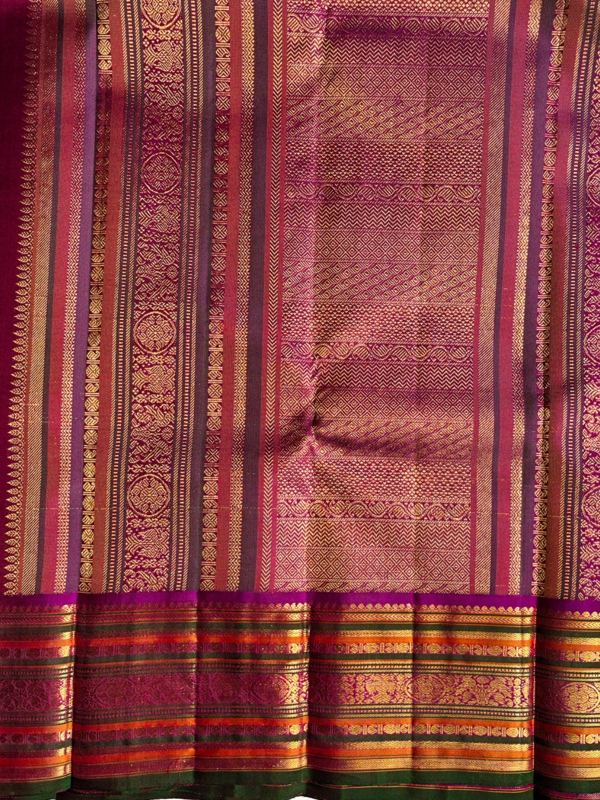 A close-up of a richly patterned silk saree body with a wide border, featuring metallic gold zari work, stripes of magenta, pink, orange, and green, and a deep magenta border.