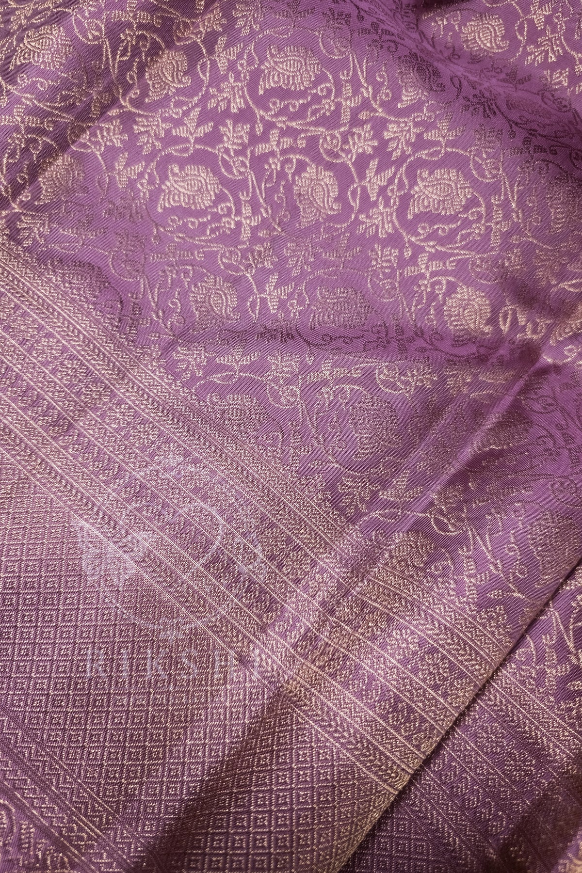 A close-up of a purple silk fabric with intricate gold zari brocade patterns and a detailed border.