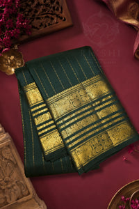 A folded dark green silk saree with intricate gold zari woven patterns, displayed on a deep maroon background with traditional decorative props.