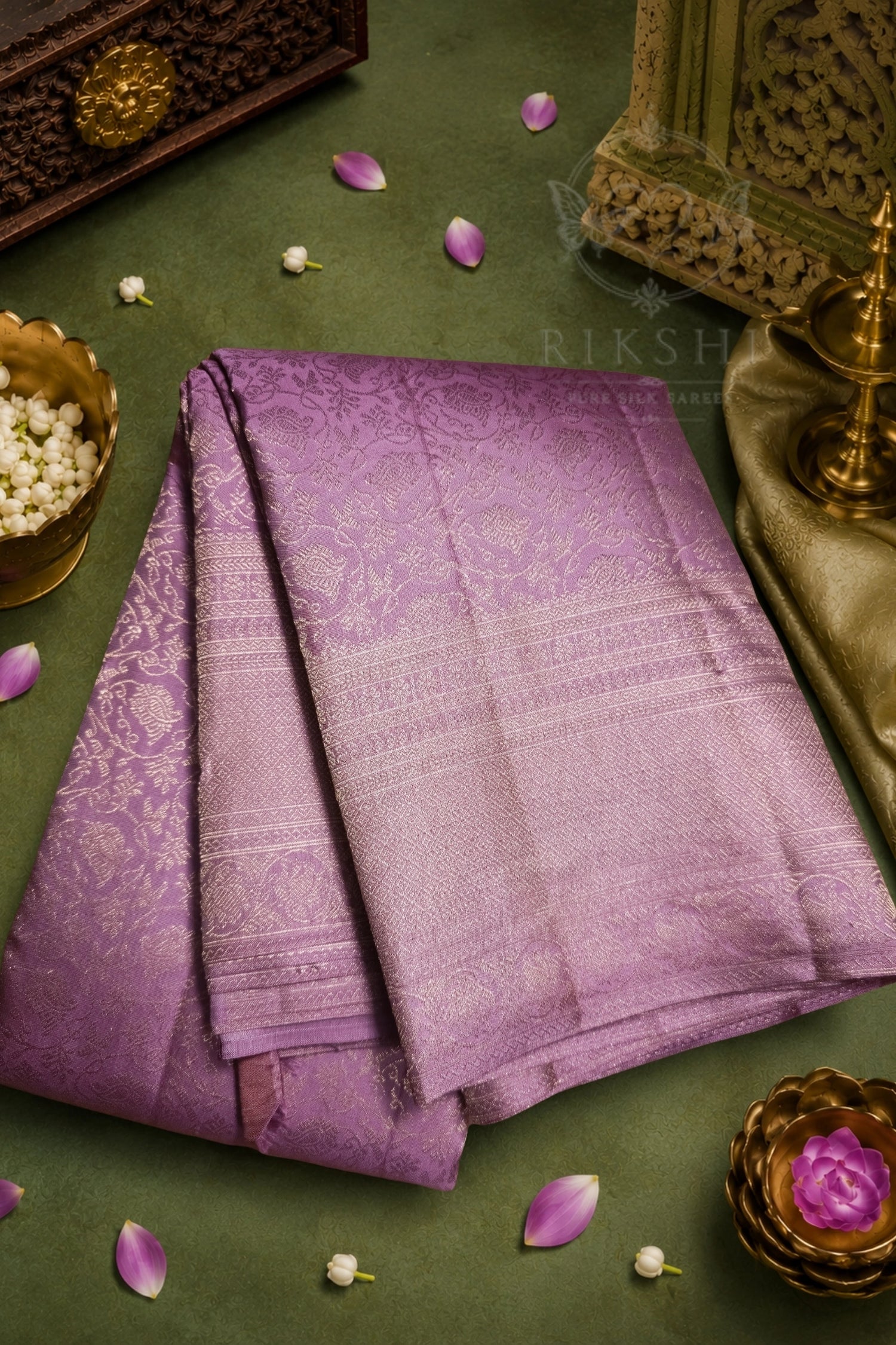 A folded purple silk saree with gold zari embroidery, displayed on a textured olive green surface with decorative props including a brass lamp, jasmine flowers, and wooden ornaments.