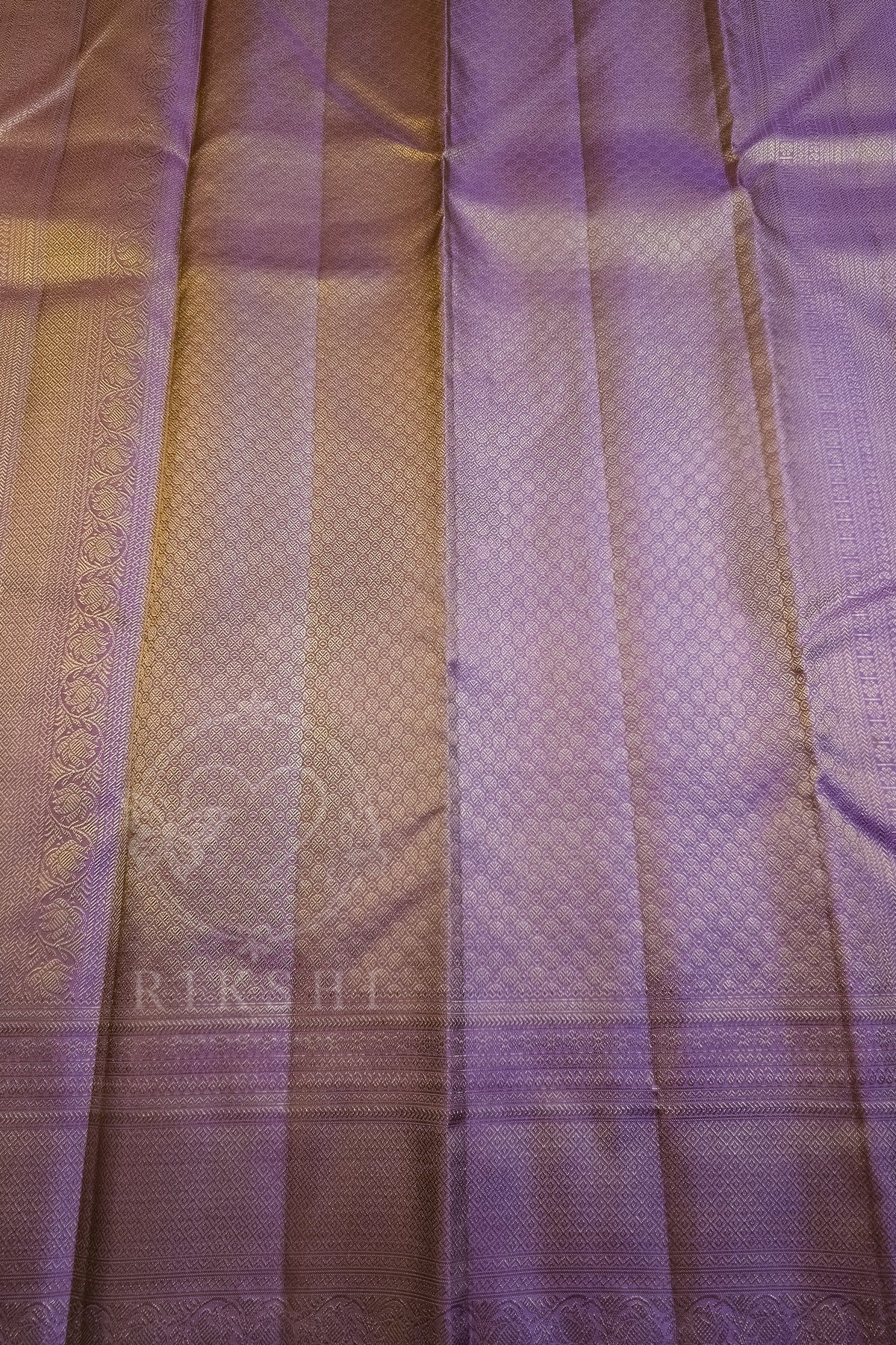 A close-up of a purple silk saree fabric with a subtle woven pattern, pleated texture, and a decorative border. A faint watermark logo 'RIKSHI' is visible near the bottom left.