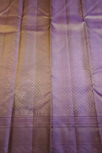 A close-up of a purple silk saree fabric with a subtle woven pattern, pleated texture, and a decorative border. A faint watermark logo 'RIKSHI' is visible near the bottom left.