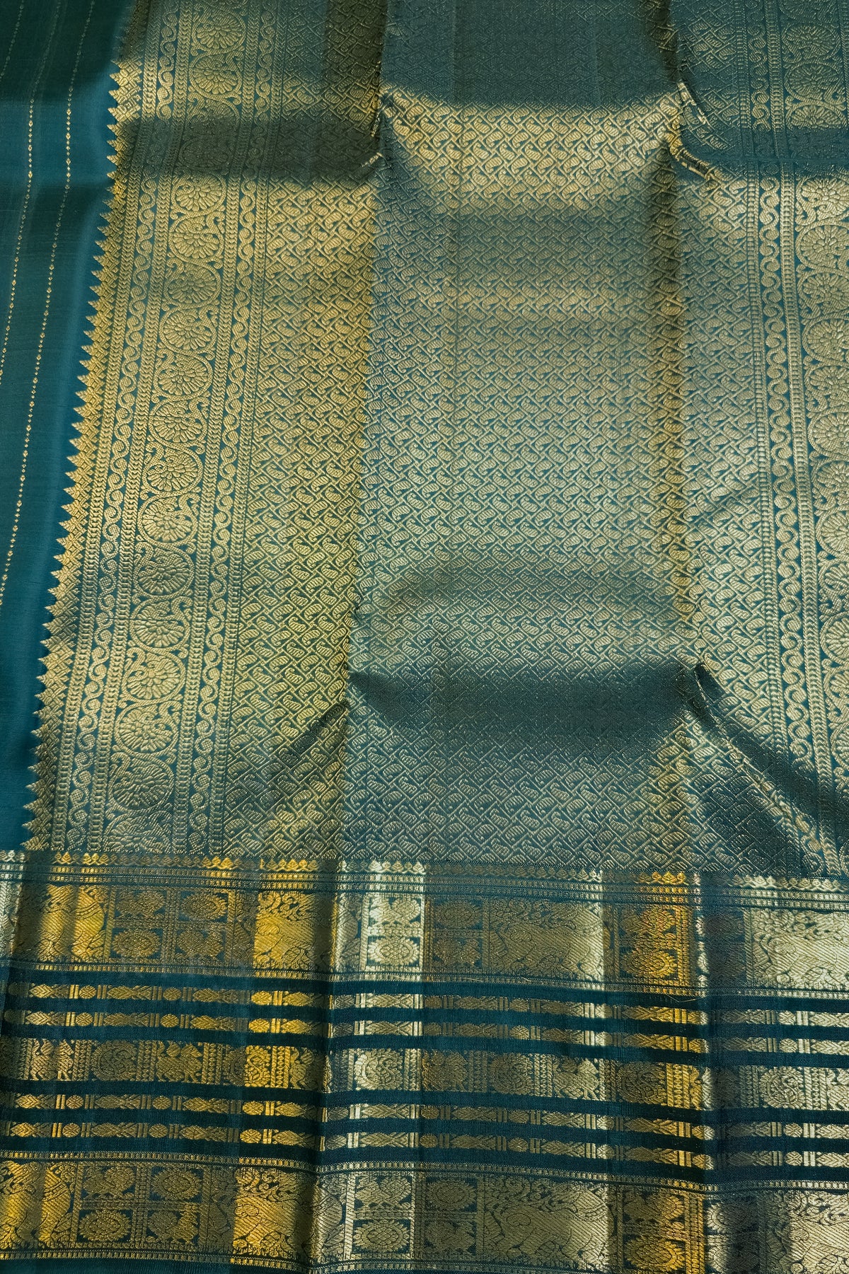 A close-up of a traditional Indian silk saree fabric, featuring a teal blue base with intricate gold zari woven patterns and a wide decorative border.