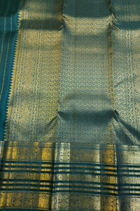 A close-up of a traditional Indian silk saree fabric, featuring a teal blue base with intricate gold zari woven patterns and a wide decorative border.