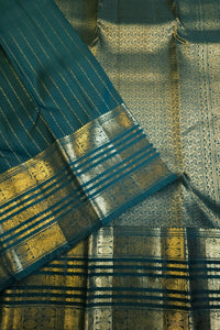 A close-up of a teal blue and gold patterned fabric, likely a saree, showing intricate metallic zari work and woven details.