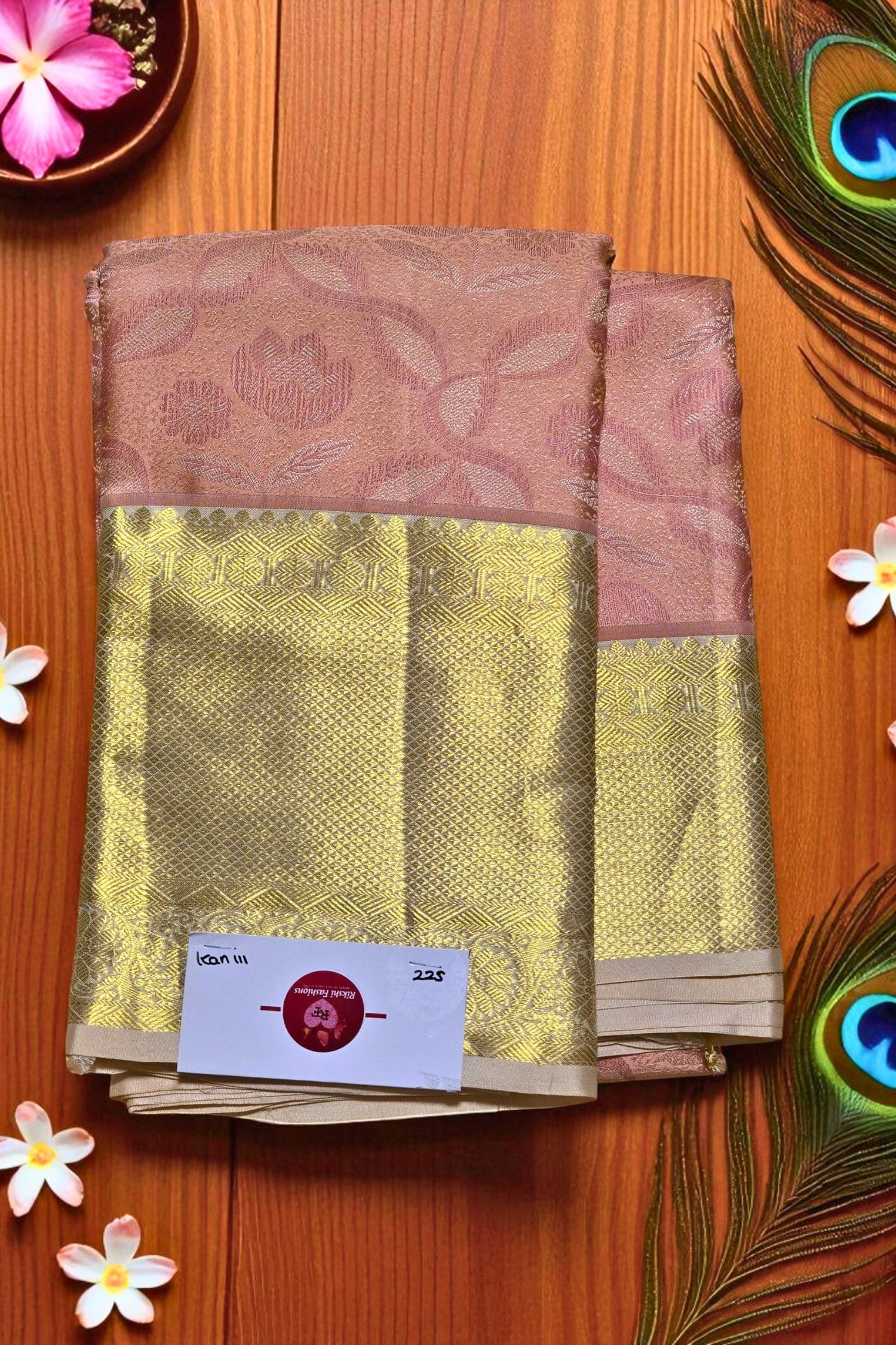 Saree with a pink and gold pattern on a wooden surface with flowers and peacock feathers.
