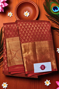 Red and gold saree with a brand label on a wooden surface with flowers and a peacock feather.