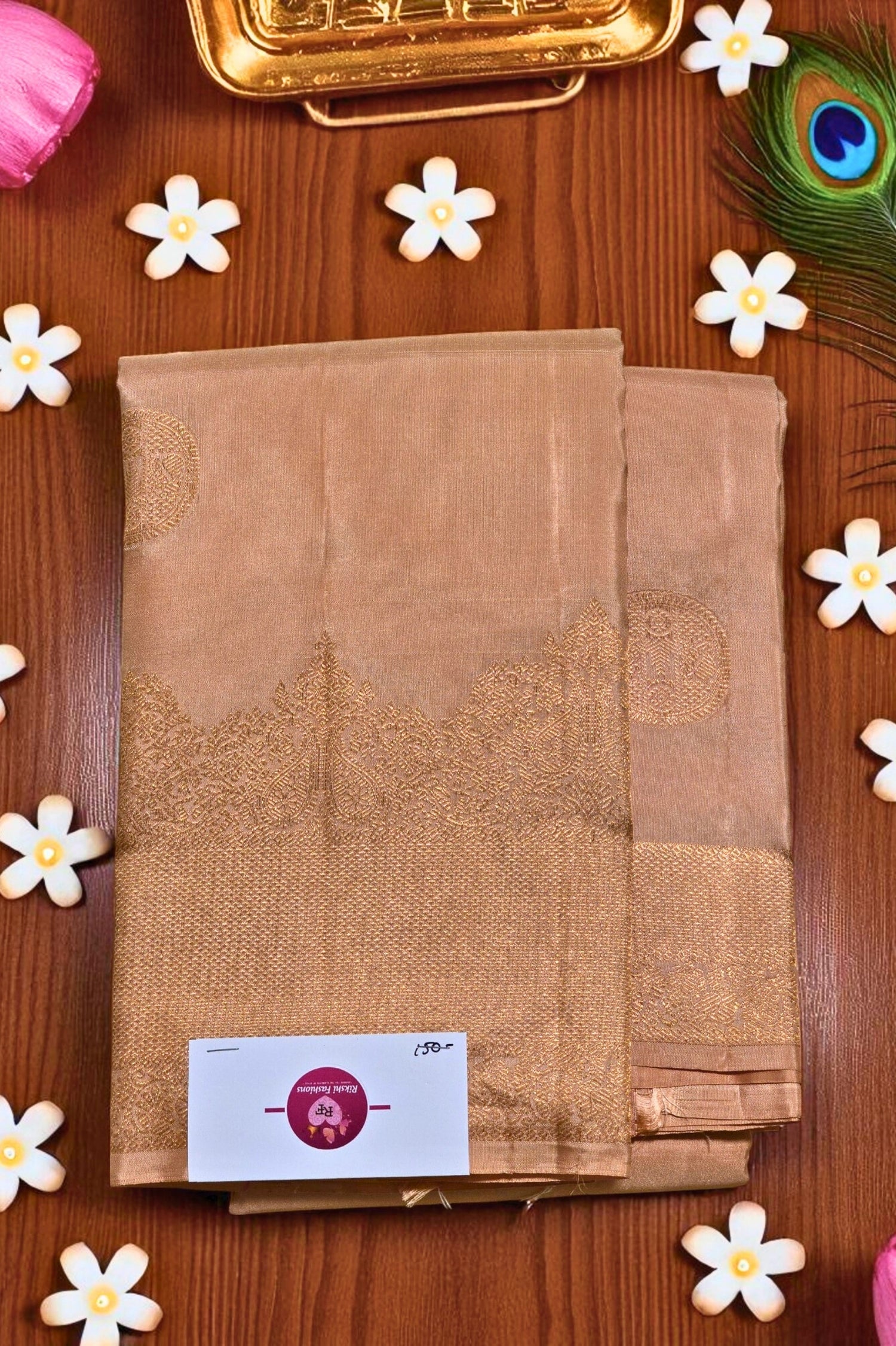 Brown saree with a brand label on a wooden surface with decorative flowers and a peacock feather.