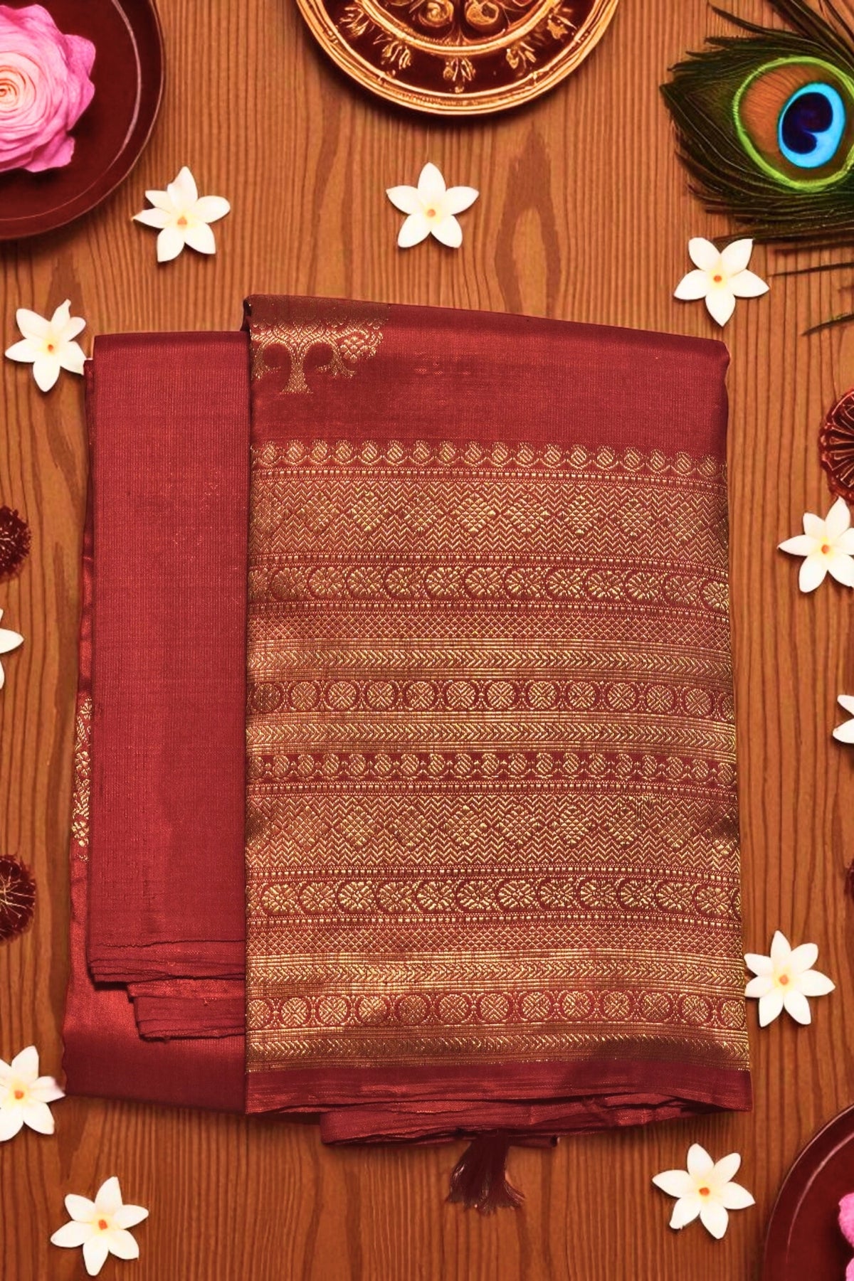 Maroon saree with gold patterns on a wooden background with flowers and a peacock feather.
