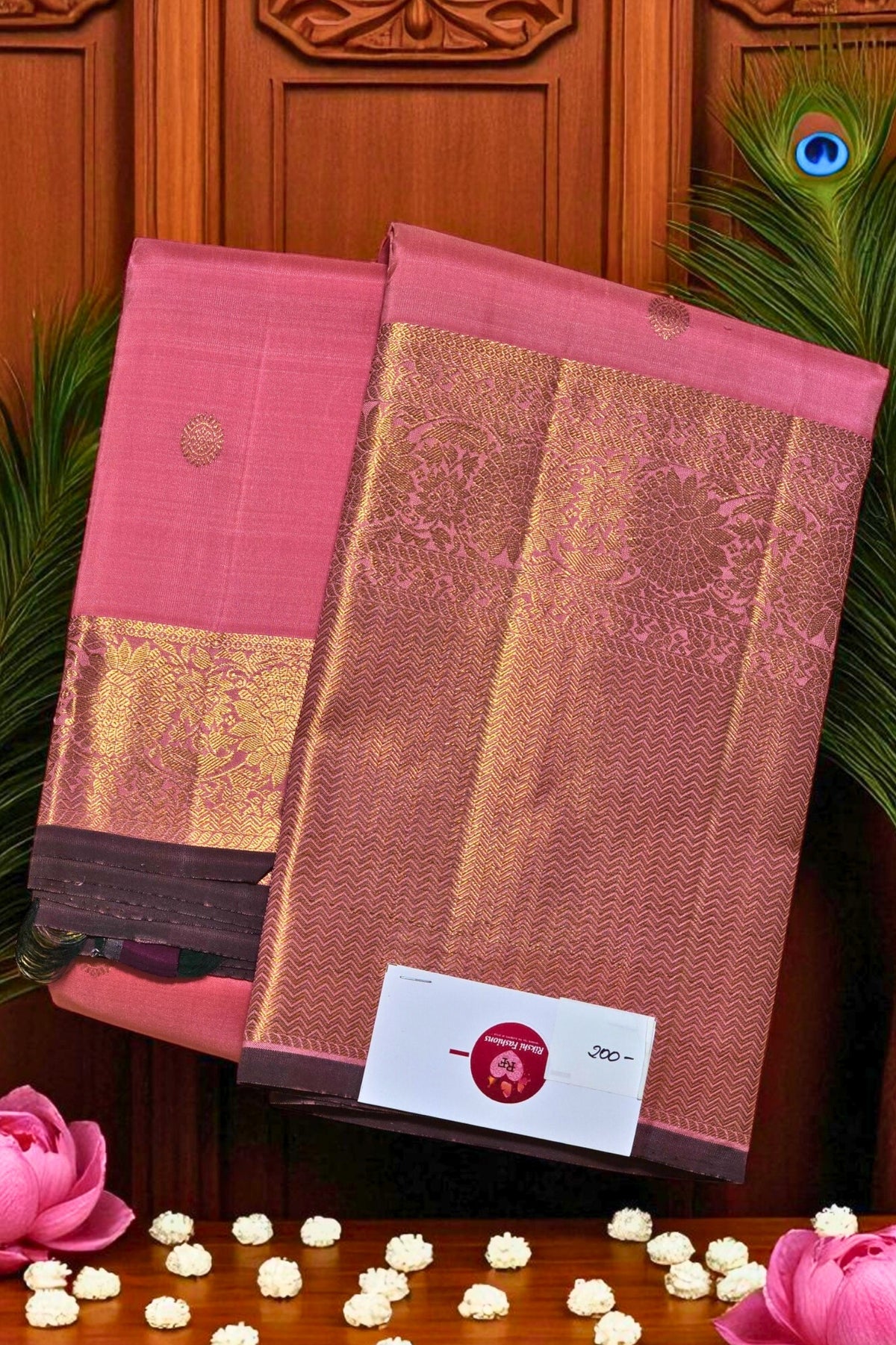Two pink sarees with gold patterns on a wooden background with peacock feathers and flowers.