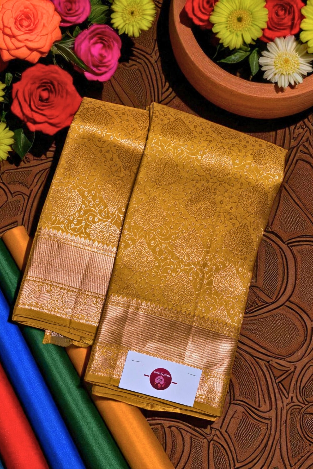 Gold saree with a brand label on a patterned surface with flowers and colorful fabrics.