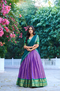 Woman in a green and purple saree standing in a garden with pink flowers and greenery.