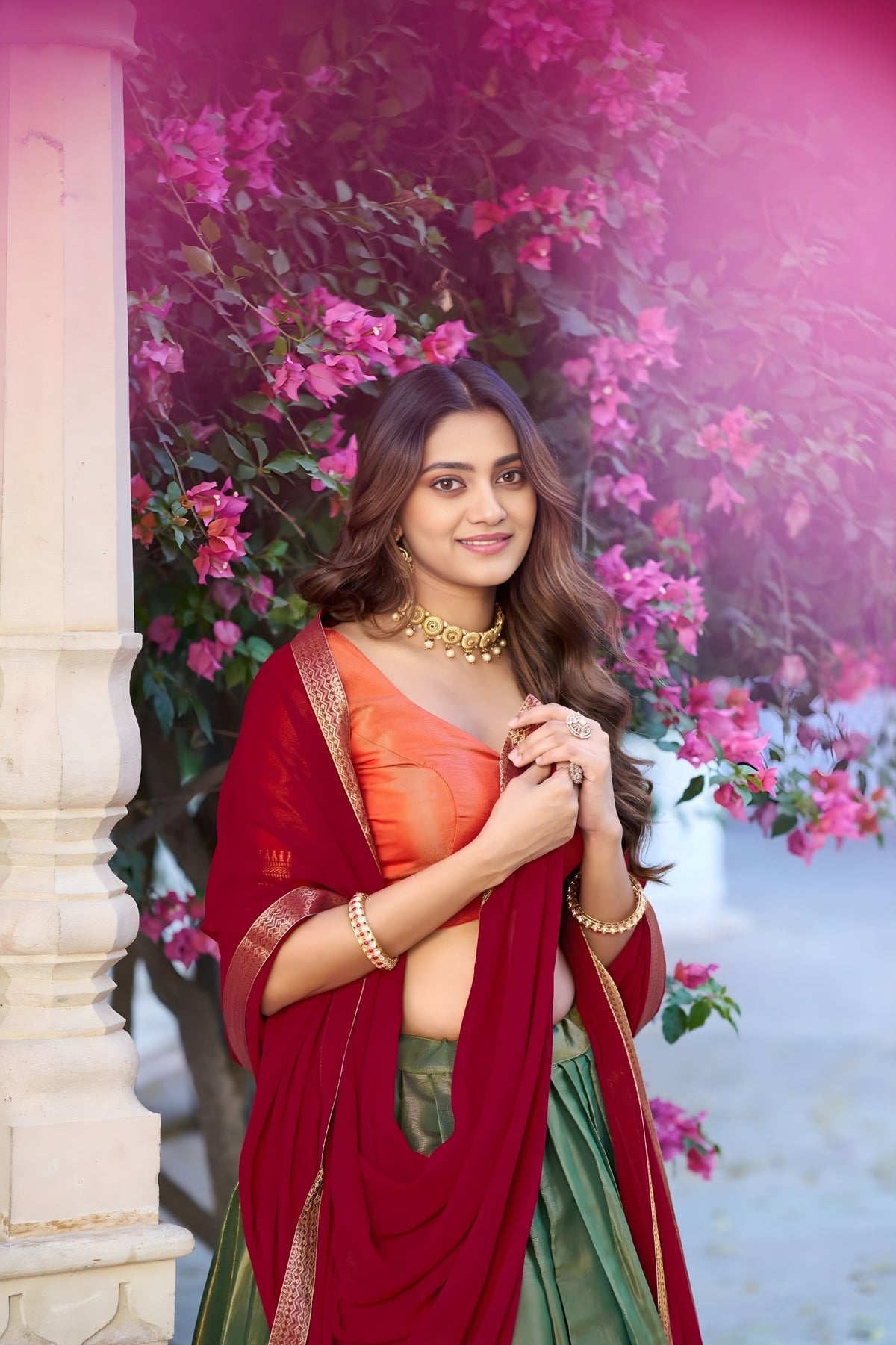 Woman in a red and green saree standing in front of pink flowers